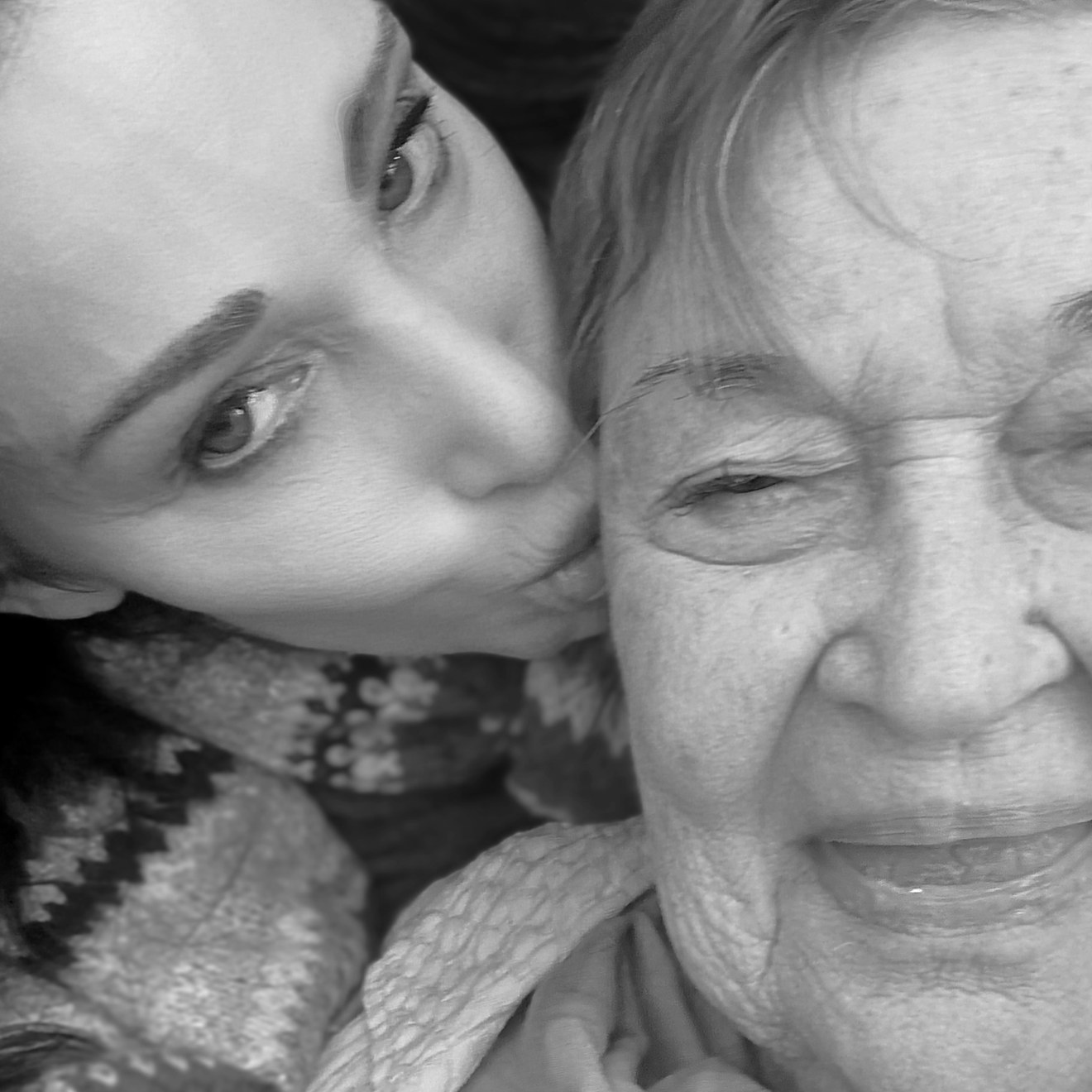 A close portrait of two women cheek to cheek: the younger one kissing the older woman's cheek, the older woman looking into the camera, smiling, in a soft scarf.