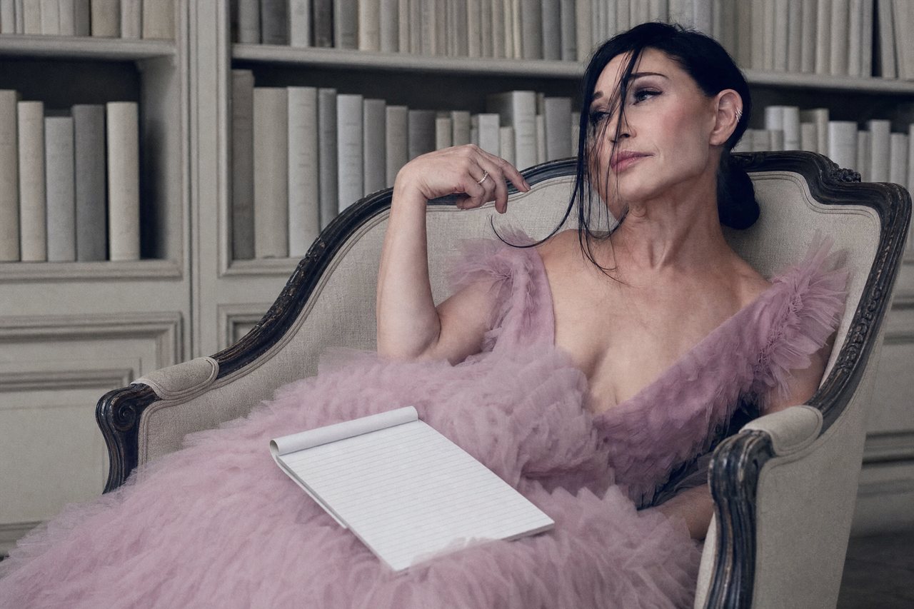 A dark-haired woman in a pink tulle gown reclining on an antique settee in a library, a blank legal pad open across her lap.